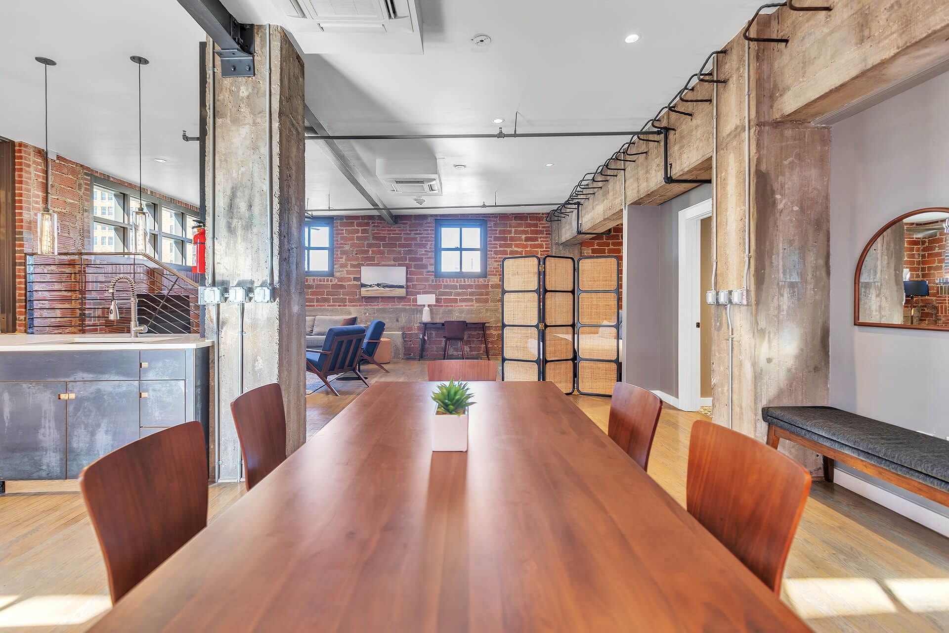 A spacious room with a long wooden table, several chairs, a small plant, and a brick wall with a window.