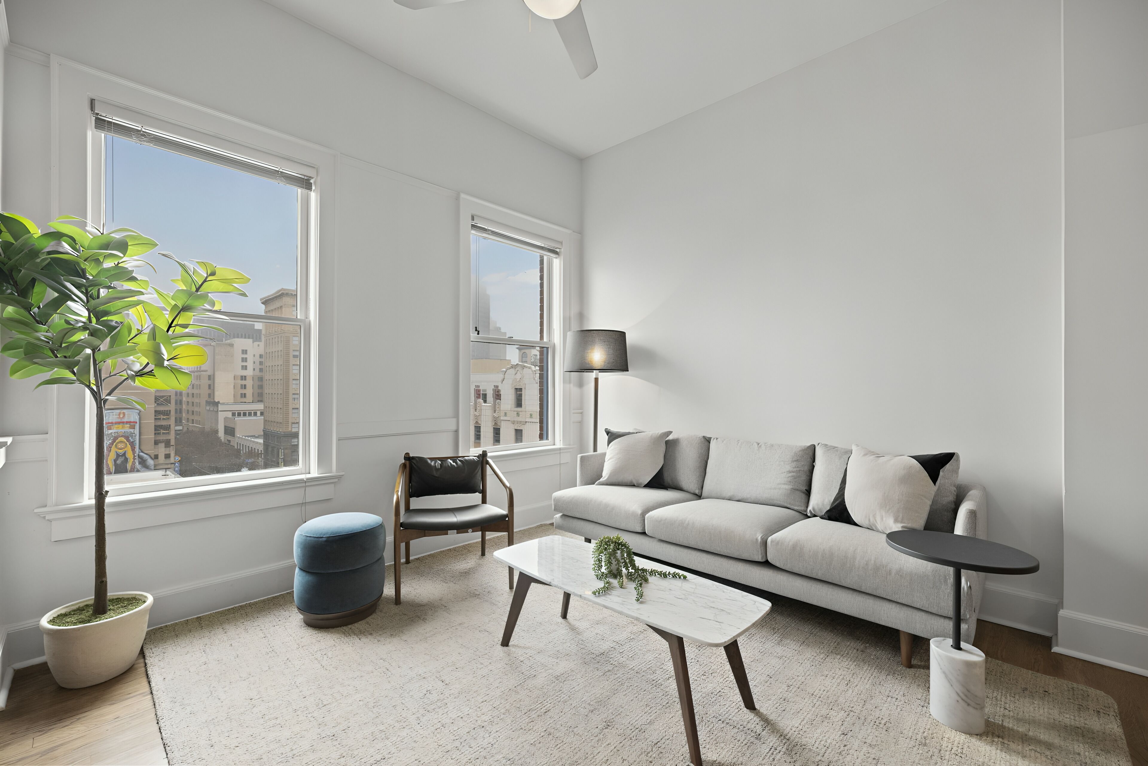A modern living room with a sofa, coffee table, and chair, large windows, and a potted plant.