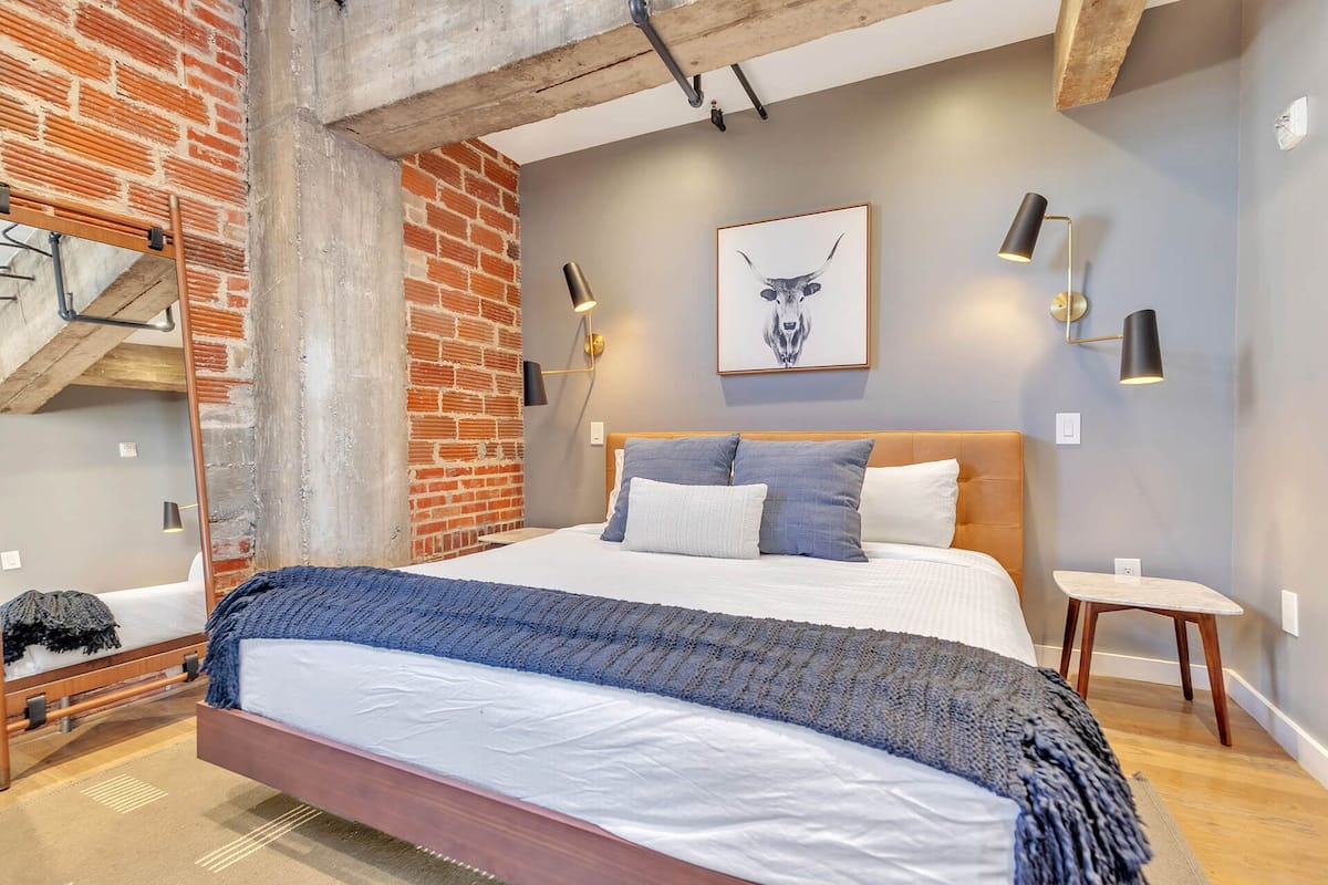 Un dormitorio con pared de ladrillo, cabecero de madera, cama con ropa de cama blanca y azul, manta azul y dos lámparas fijadas a la pared.