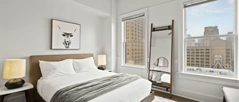 Quarto de hotel moderno com uma cama grande, uma mesa de cabeceira com abajur, uma janela com vista para a cidade e uma obra de arte emoldurada na parede.