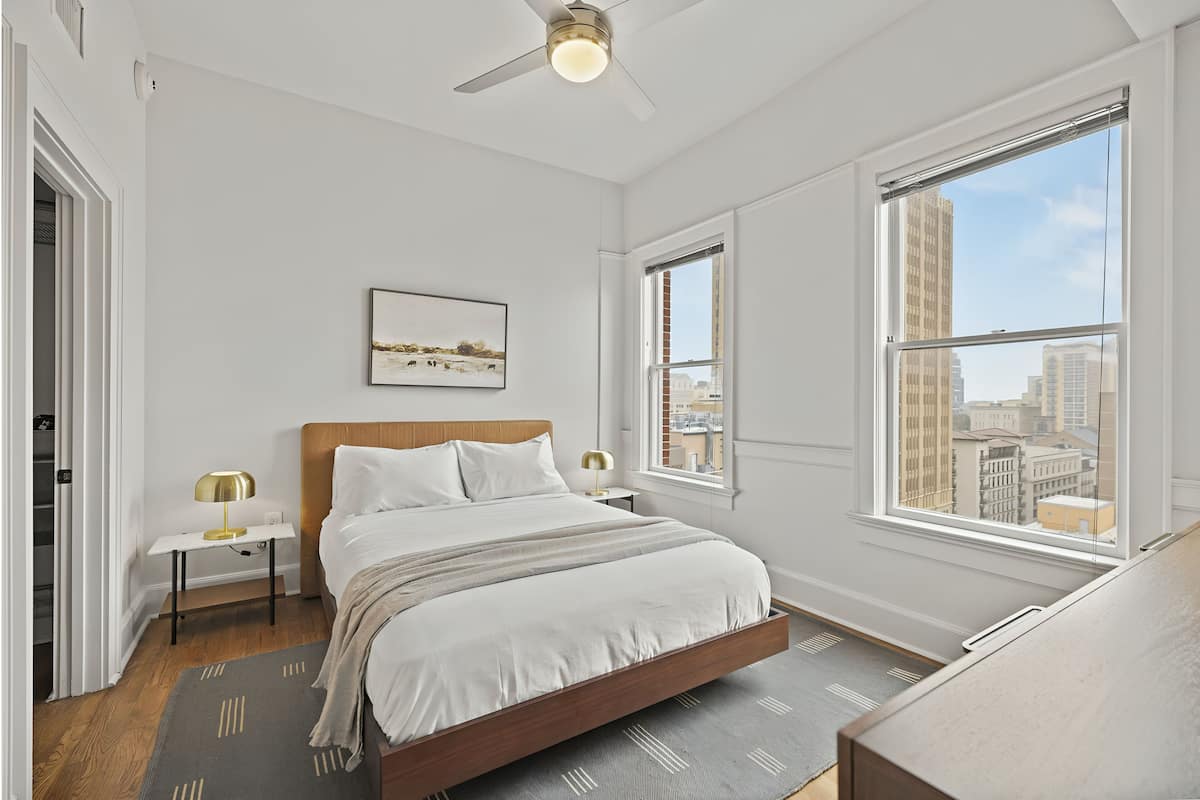 Un dormitorio con cama, mesita de noche, lámpara, ventana con vista a la ciudad y ventilador de techo.