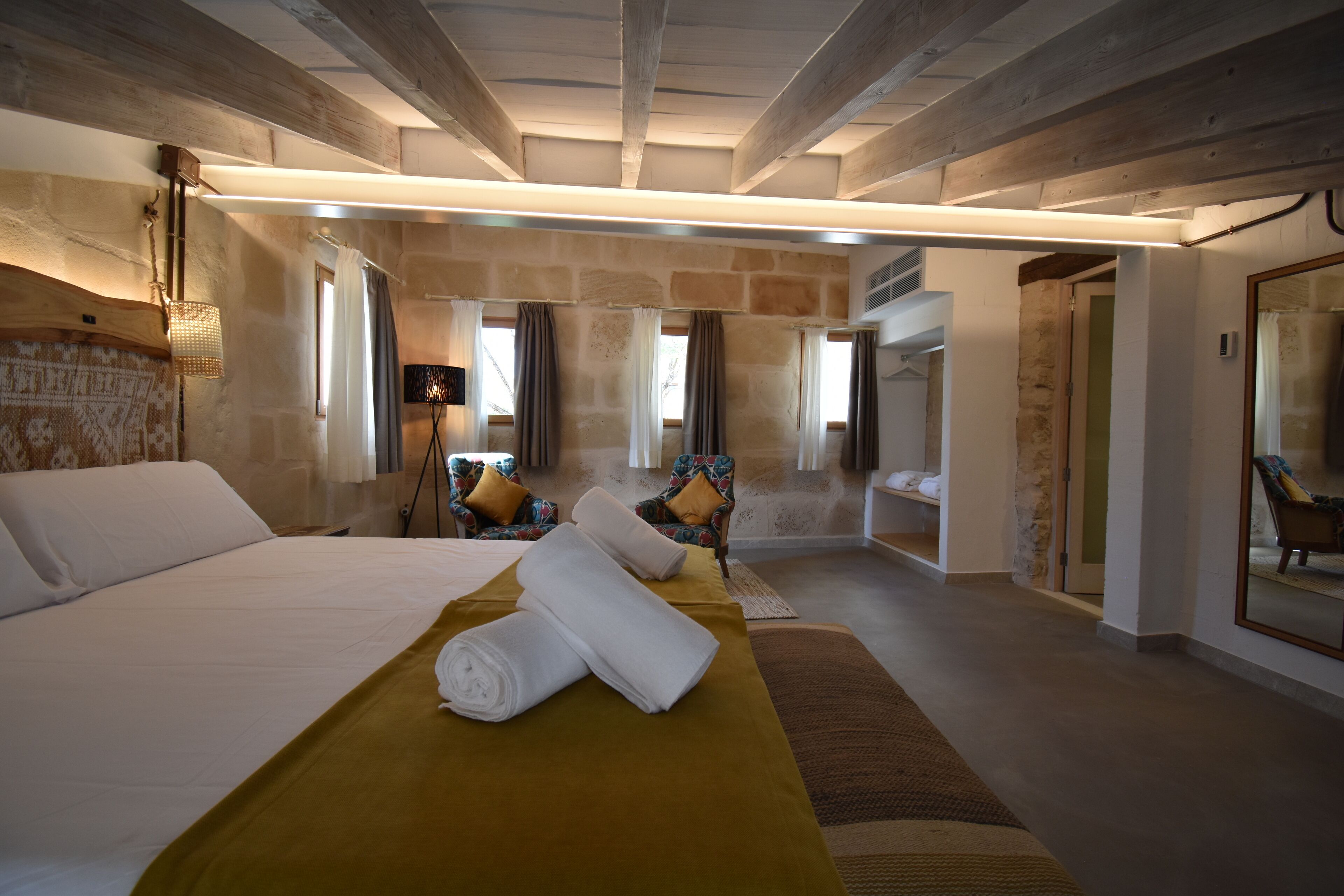 Una habitación de hotel moderna con una cama grande, un espacio de descanso con sofá y sillones, un baño con espejo y una pared de piedra.