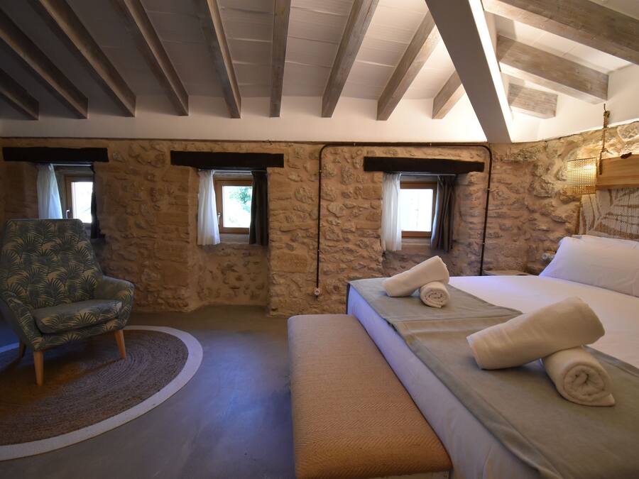A bedroom with a stone wall, a bed with rolled towels, a chair, and a window with curtains.