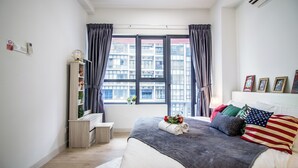 A bedroom with a large window, a bed with multiple pillows, a bedside table, and a small shelf with plants. - Arte plus KLCC By Tenang Home (Kuala Lumpur)