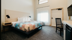 Habitación de hotel con cama, escritorio, silla, mesita de noche, lámpara, ventana con cortinas y puerta.