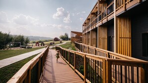 Una terraza de madera que conduce a un edificio con balcones, un espacio con césped y un granero rojo al fondo.