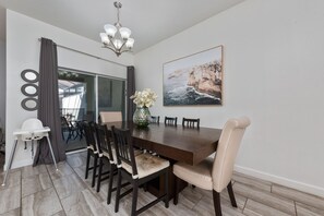 Uma sala de jantar com uma grande mesa de madeira, seis cadeiras, um vaso com flores e uma pintura de falésias na parede.