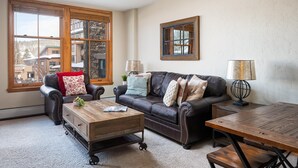A cozy living room with a leather sofa, a wooden coffee table, and a large window offering a view of buildings and trees. - Ski In Ski Out | Zephyr Mountain Lodge 2219 | Hot Tub | Base Area (Winter Park)