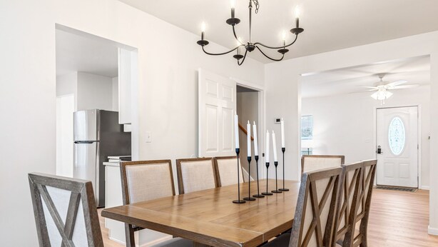 A dining area with a wooden table, six chairs, a chandelier, and a modern kitchen in the background. - Renovated 5BR | Group Space + Large Yard (Wentzville)
