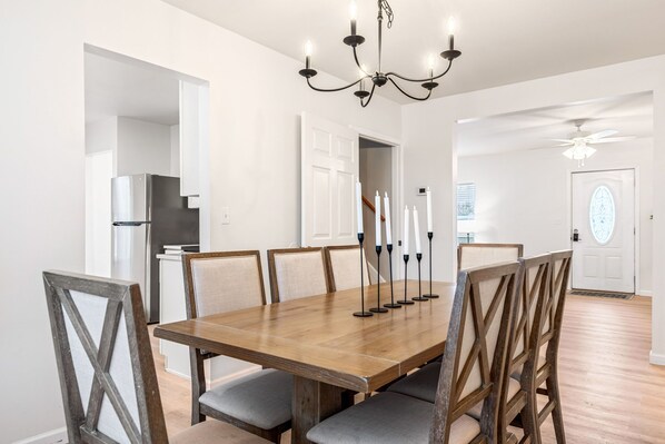 A dining area with a wooden table, six chairs, a chandelier, and a modern kitchen in the background. - Renovated 5BR | Group Space + Large Yard (Wentzville)