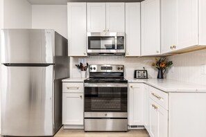 A modern kitchen with white cabinetry, stainless steel appliances including a refrigerator, microwave, and oven, and a countertop with a coffee maker and utensils. - Renovated 5BR | Group Space + Large Yard (Wentzville)