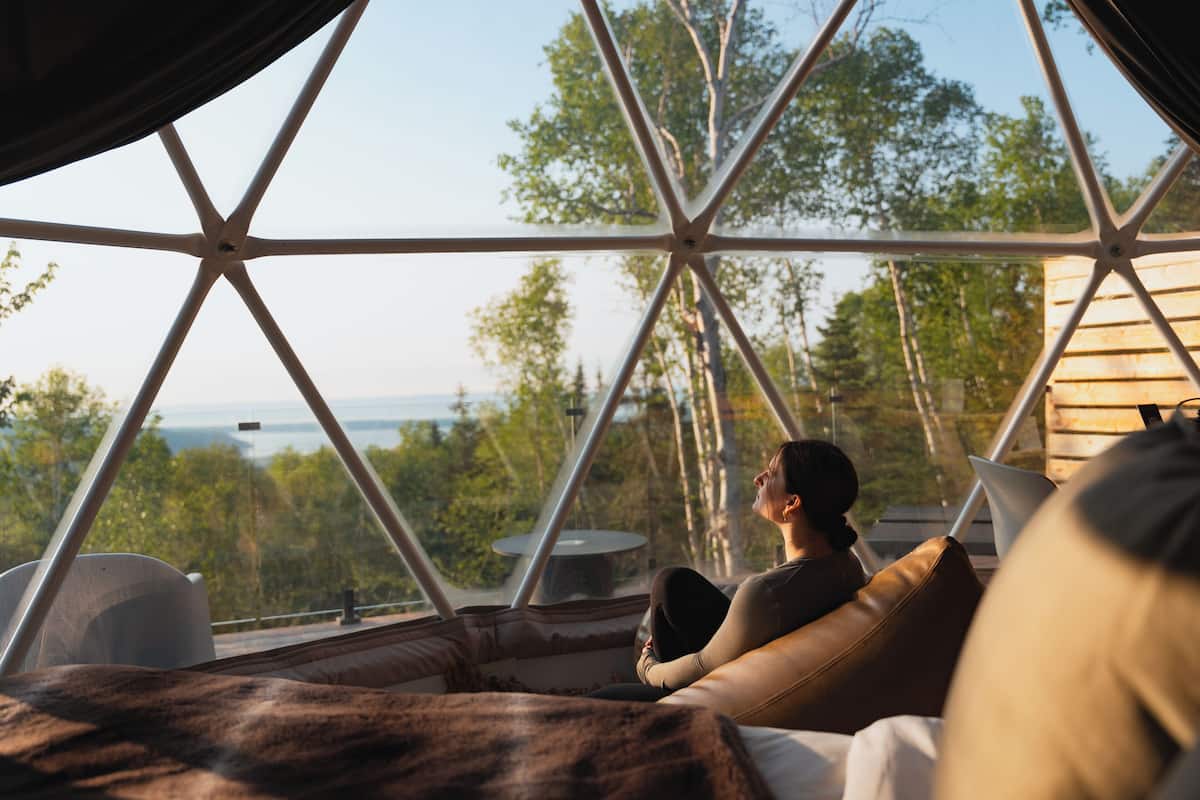 Una habitación con un ventanal, vista a árboles, y una persona sentada en un sofá.