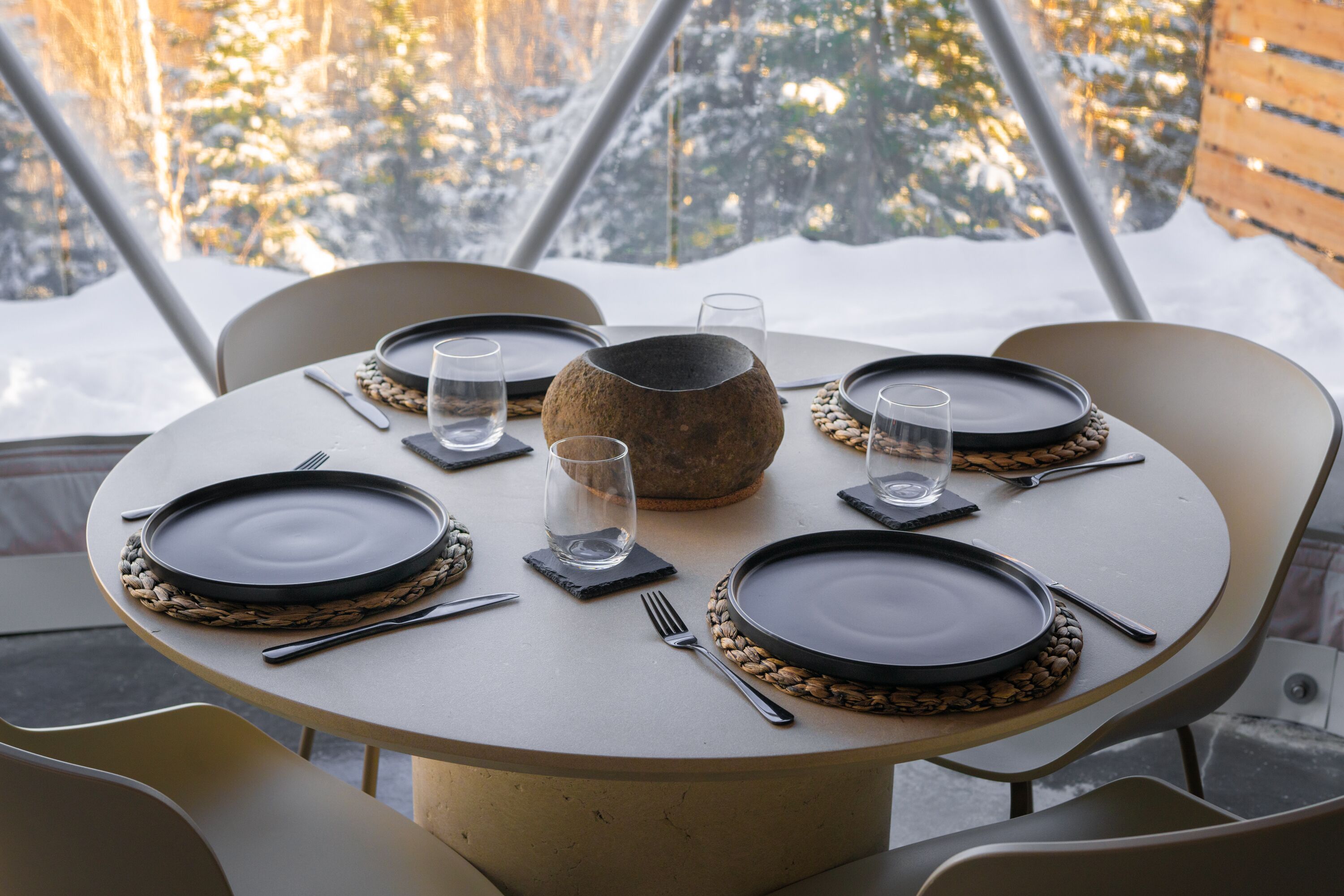 Une table de salle à manger ronde, dressée pour quatre personnes, avec des assiettes noires, des couverts et des sets de table en fibres tressées.