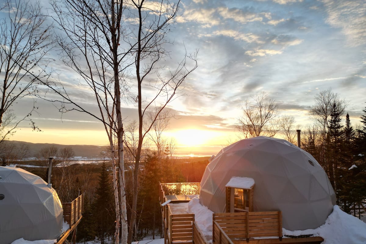 Un espacio al aire libre cubierto de nieve, con bancos de madera y una estructura abovedada, probablemente parte de un complejo turístico o establecimiento de alojamiento.