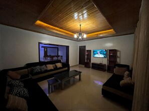 Un salon avec un canapé de couleur sombre, une table basse et un téléviseur fixé au mur. Le plafond est en bois et agrémenté d’un lustre.