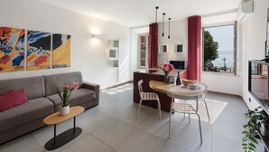 Ein modernes Wohnzimmer mit einer Couch, einem Esstisch mit Stühlen, einem kleinen Beistelltisch mit Pflanze und Meerblick durch große Fenster.