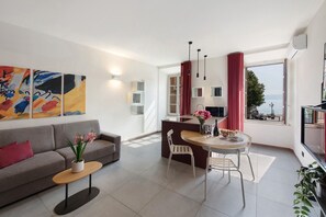 A modern living room with a sofa, a dining table with chairs, a small side table with a plant, and a view of the sea through large windows.