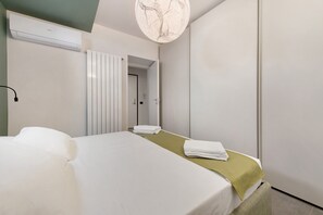 Quarto de hotel com uma cama, uma toalha verde, um radiador branco e uma porta que dá para outro quarto.