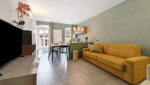 Un salon moderne comprenant un canapé, un téléviseur à écran plat, un coin repas avec des chaises et une table, ainsi qu’un coin cuisine avec un comptoir et des luminaires suspendus.