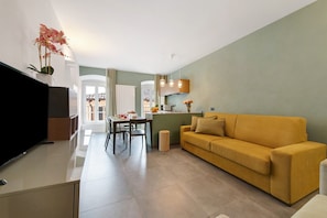 Uma sala de estar moderna com sofá, televisão de tela plana, área de jantar com cadeiras e mesa, e cozinha com bancada e luminárias pendentes.