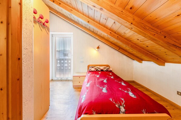 Une chambre avec un plafond en pente, un lit recouvert d’une couverture rouge, une porte en bois et une fenêtre donnant sur un beau paysage.