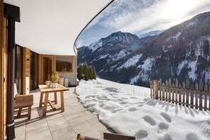 Eine Holzdiele mit Tisch und Stühlen, ein Holzzaun und Blick auf schneebedeckte Berge.