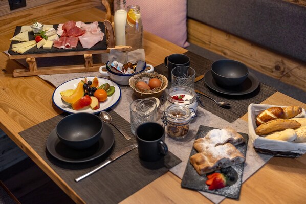 Une table dressée pour le petit-déjeuner avec des assiettes de fruits, de fromage et de charcuterie, des bols noirs et un panier de viennoiseries.