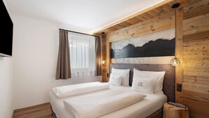 Uma cama de casal com roupa de cama branca, uma cabeceira de madeira com uma pintura de montanhas e uma mesa de cabeceira com abajur.