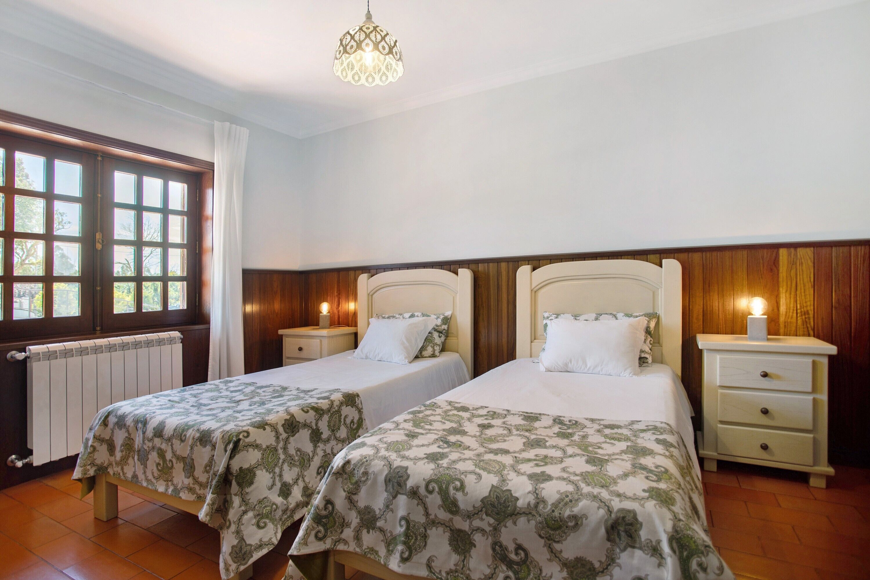 A room with two beds, a radiator, a window with curtains, and a chandelier.