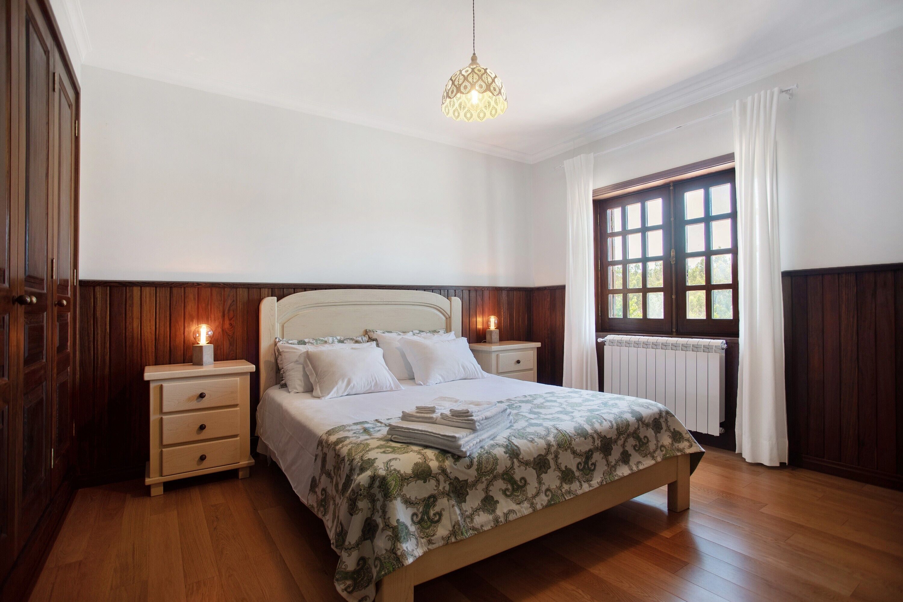 Ein Schlafzimmer mit einem hölzernen Bett, zwei Nachttischen, einem Schminktisch, einem Fenster mit Vorhängen und einer Kristallleuchter.