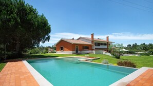 Uma piscina com deck ladrilhado, uma casa com telhado vermelho e uma grande árvore ao fundo.