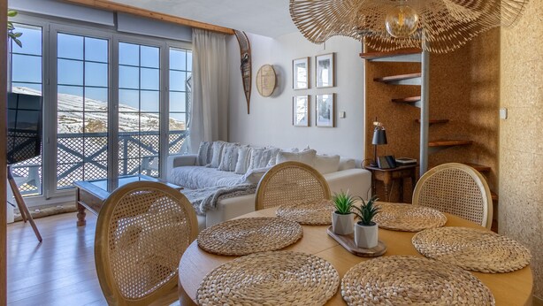 A cozy living room with a wooden dining table, wicker chairs, and a large window overlooking a snowy landscape. - Homity Dúplex Sierra Nevada Mt-3-p1-345 (Sierra Nevada)