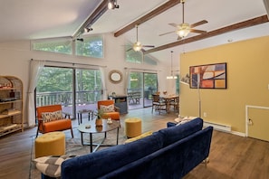 A spacious living room with a large sofa, a dining table, and a balcony with a view of trees. - Cozy Family Cabin • Steps to Lake • Canoe • Firepit • Pet Friendly (Pocono Lake)