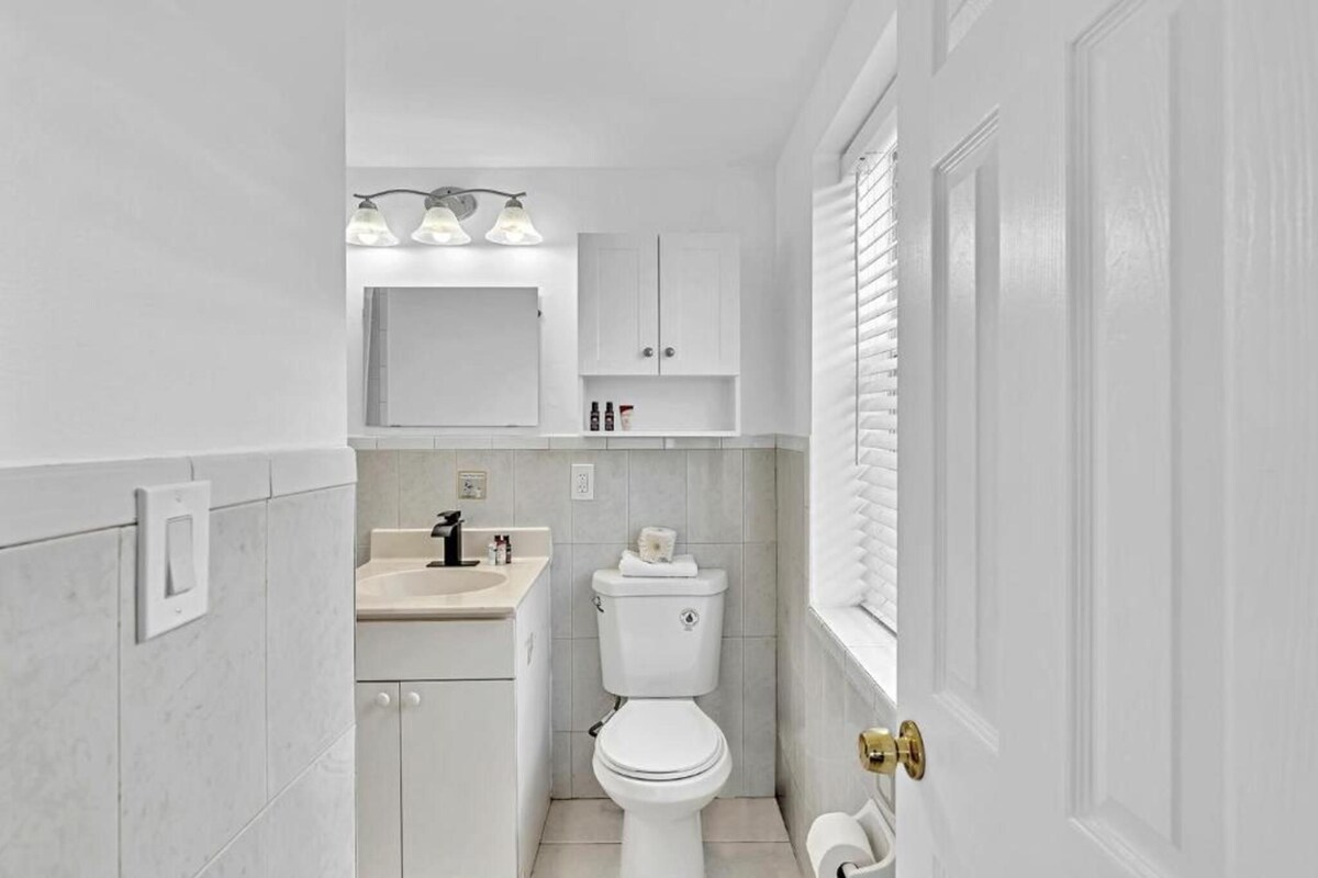 A compact bathroom with a toilet, sink, and vanity, featuring a white color scheme and a window with blinds.
