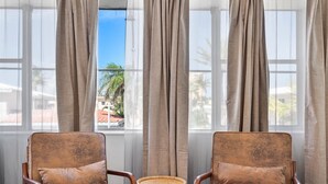 Quarto com duas poltronas marrons e uma mesa redonda pequena, janelas amplas com cortinas leves e vista para palmeiras e edifícios.