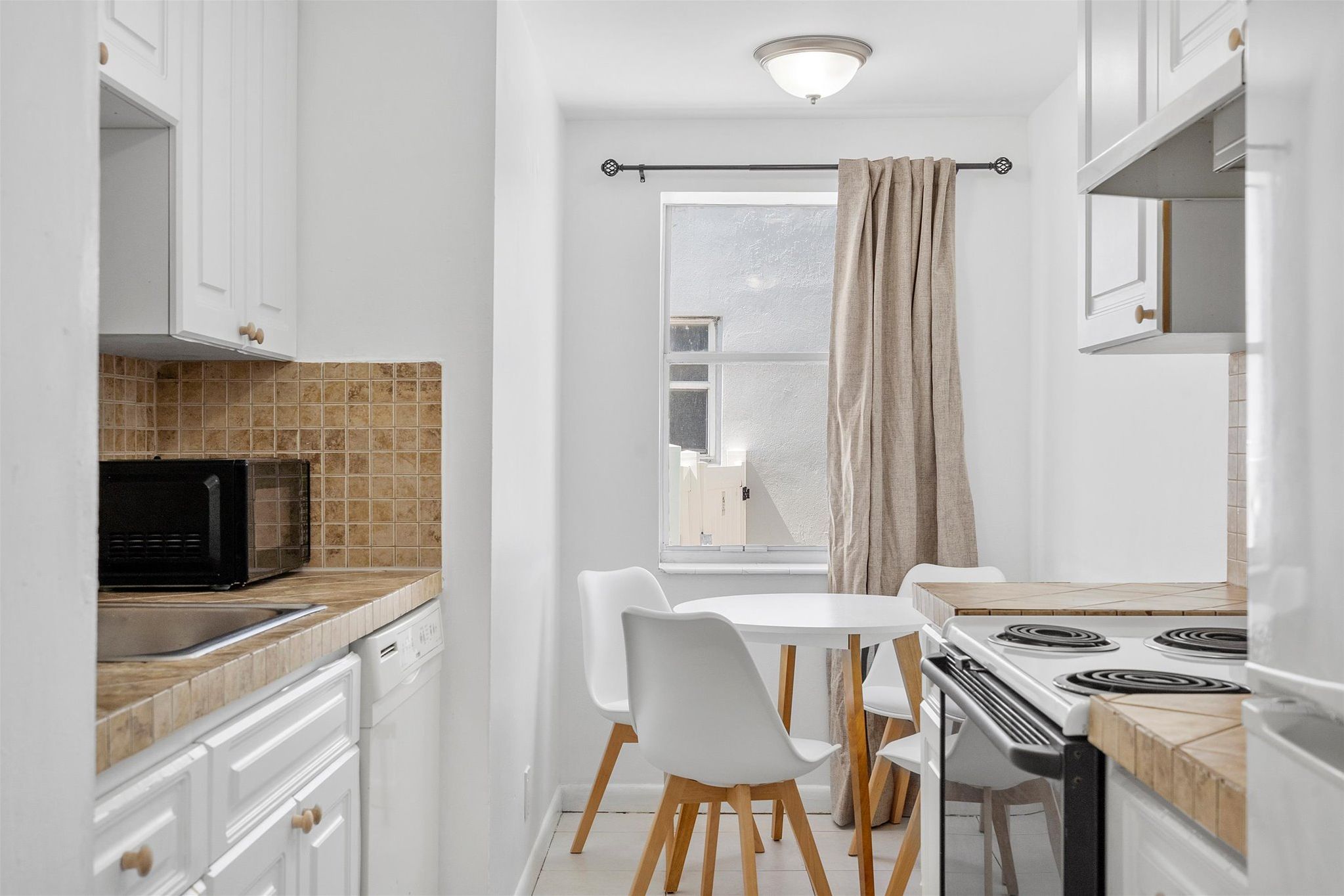 Une cuisine étroite avec des meubles blancs, un four à micro-ondes, une cuisinière et une fenêtre avec des rideaux.