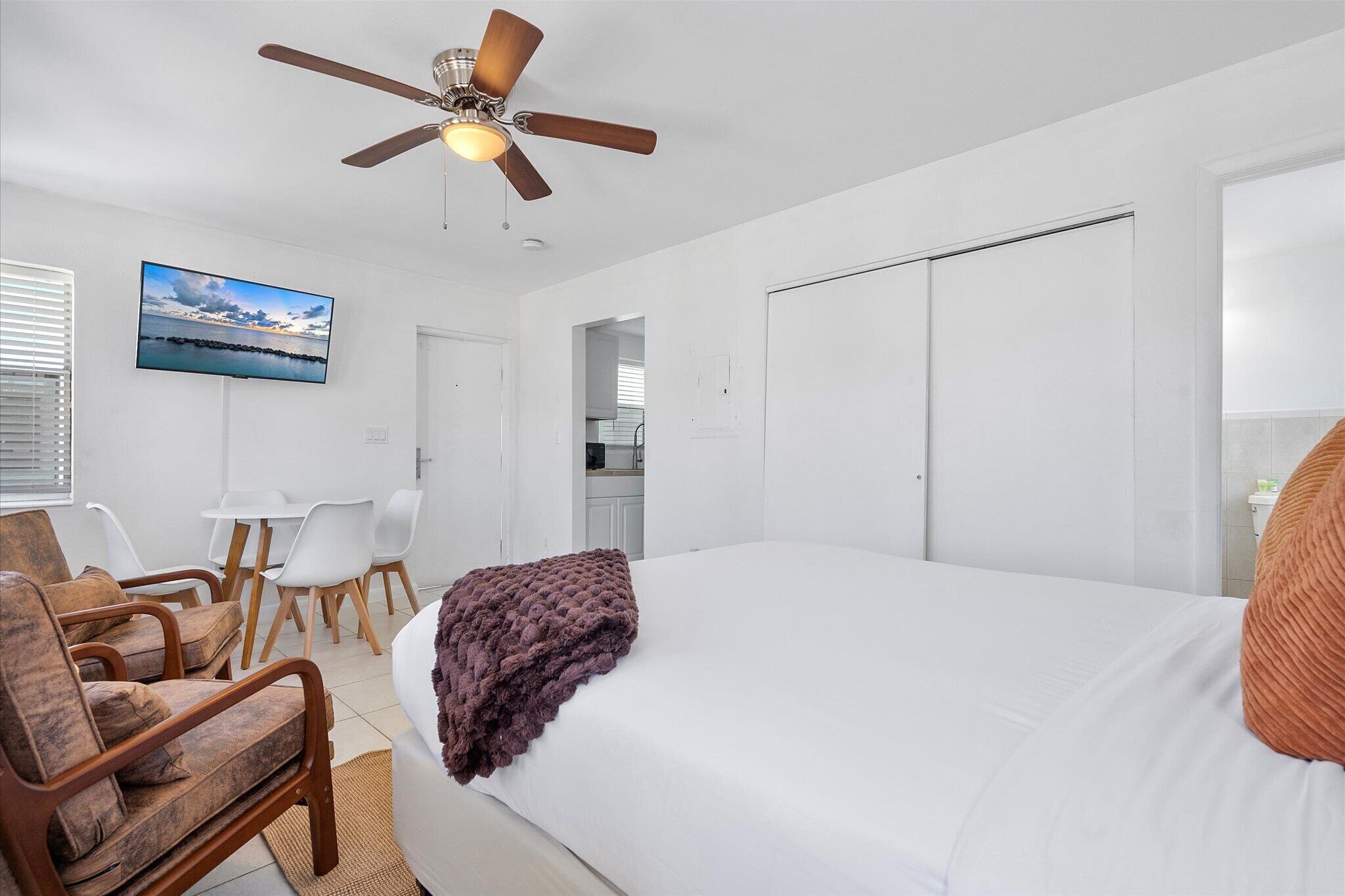 Une chambre avec un lit, un ventilateur de plafond, une télévision, un coin repas avec des chaises et, au fond, une cuisine.