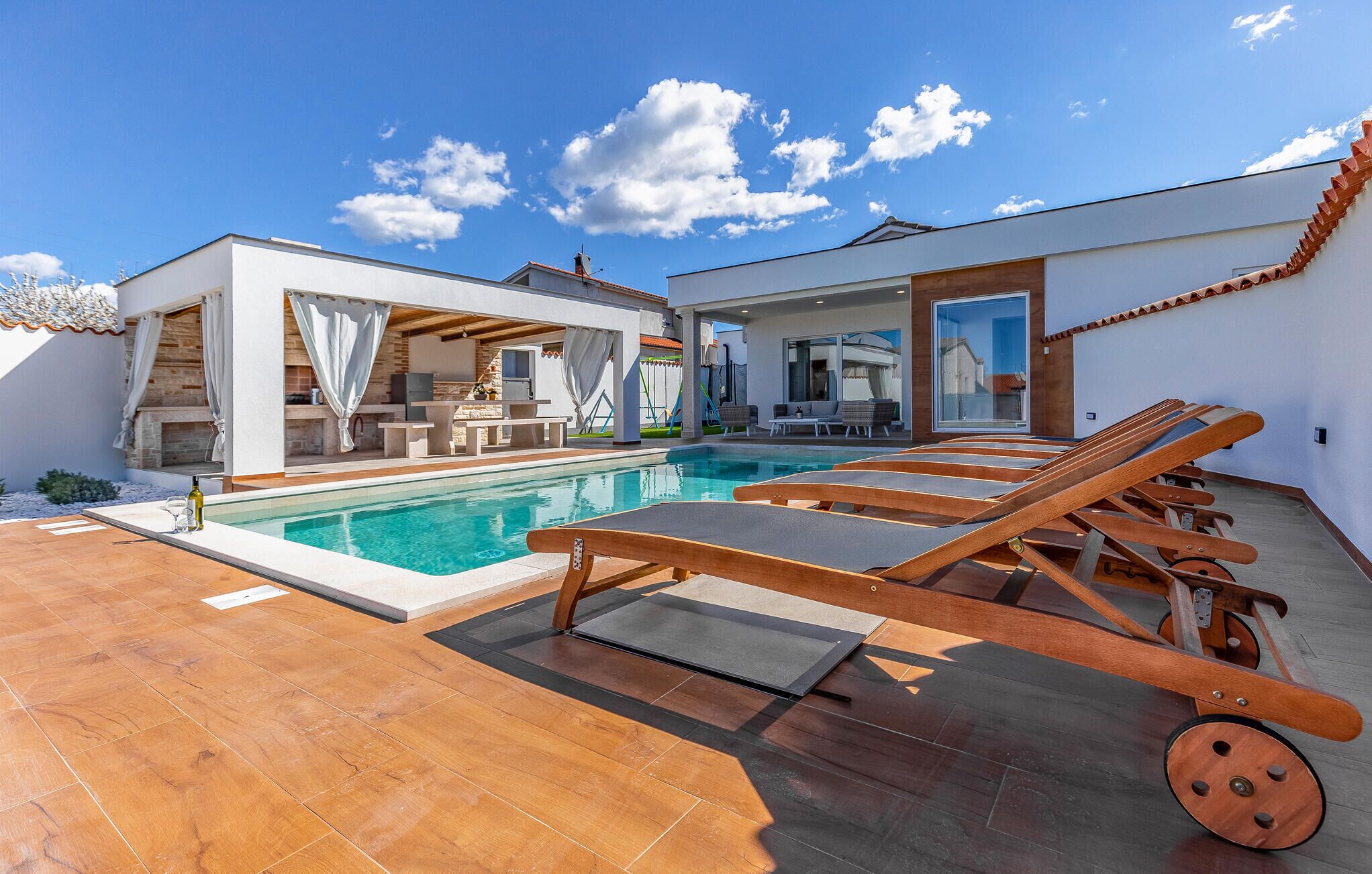 A modern outdoor pool area with lounge chairs, a barbecue setup, and a covered seating area.