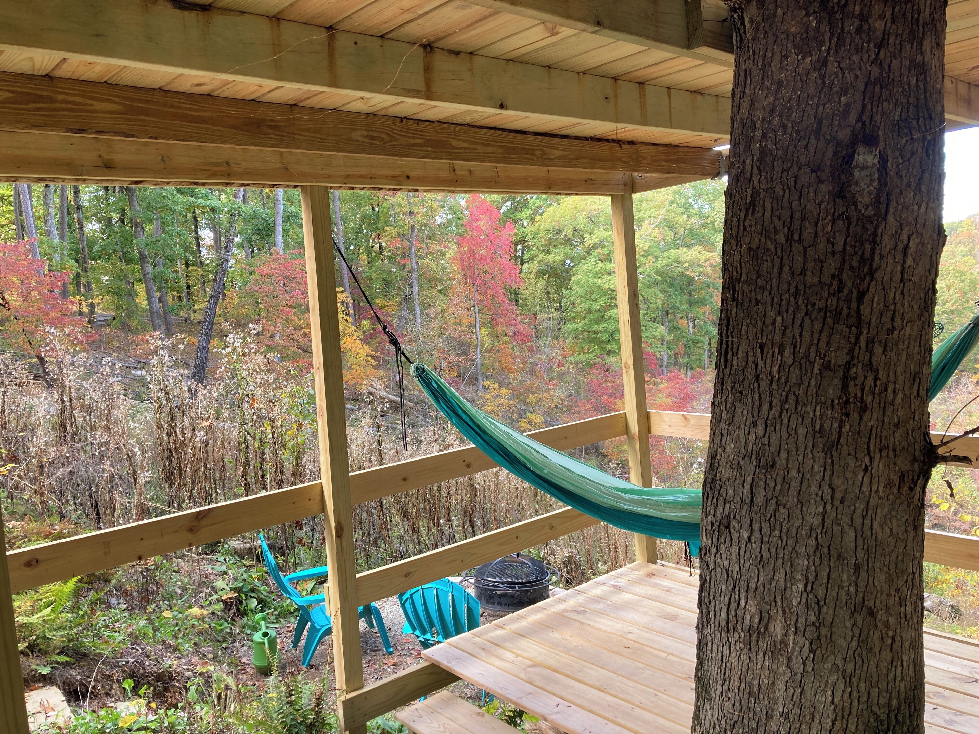 A wooden deck with a hammock, a table, and chairs, overlooking a forested area with autumn foliage.