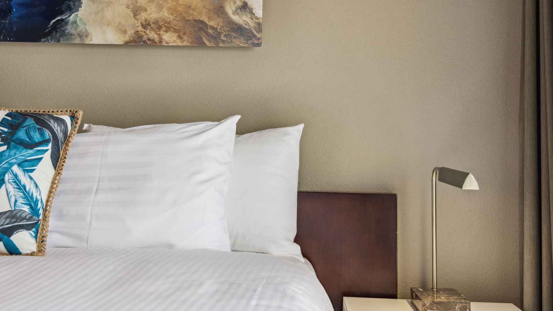 Hotelkamer met een bed, witte kussens, een sierkussen met een blauw en goud patroon, een nachtkastje met een lamp en een schilderij aan de muur.