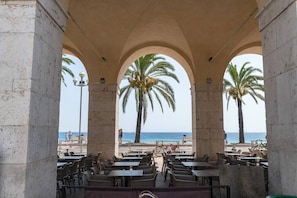 Um café ao ar livre com arcos de pedra, mesas e cadeiras, com vista para uma praia com palmeiras e o mar.