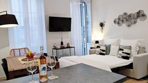 A modern hotel room with a bed, a desk with a bottle and glasses, a chair, a TV, and a window with curtains.