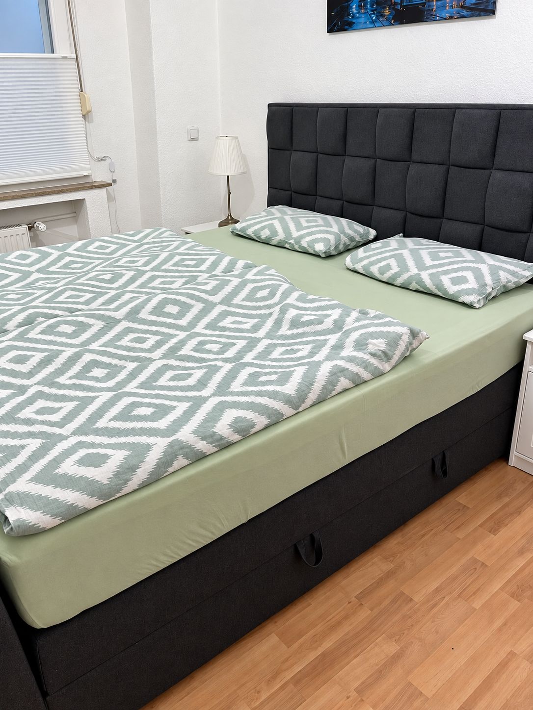 Una cama con colcha y almohadas estampadas, cabecera oscura, mesita de noche con lámpara y un cuadro colgado en la pared.
