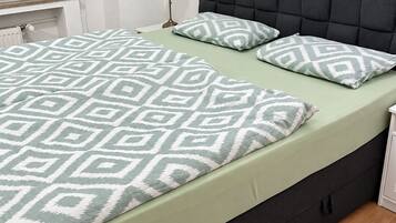 Una cama con colcha y almohadas estampadas, cabecera oscura, mesita de noche con lámpara y un cuadro colgado en la pared.