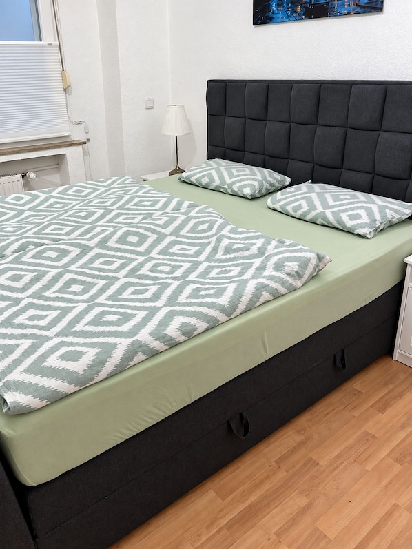 A bed with a patterned bedspread and pillows, a dark headboard, a bedside table with a lamp, and a wall-mounted picture.