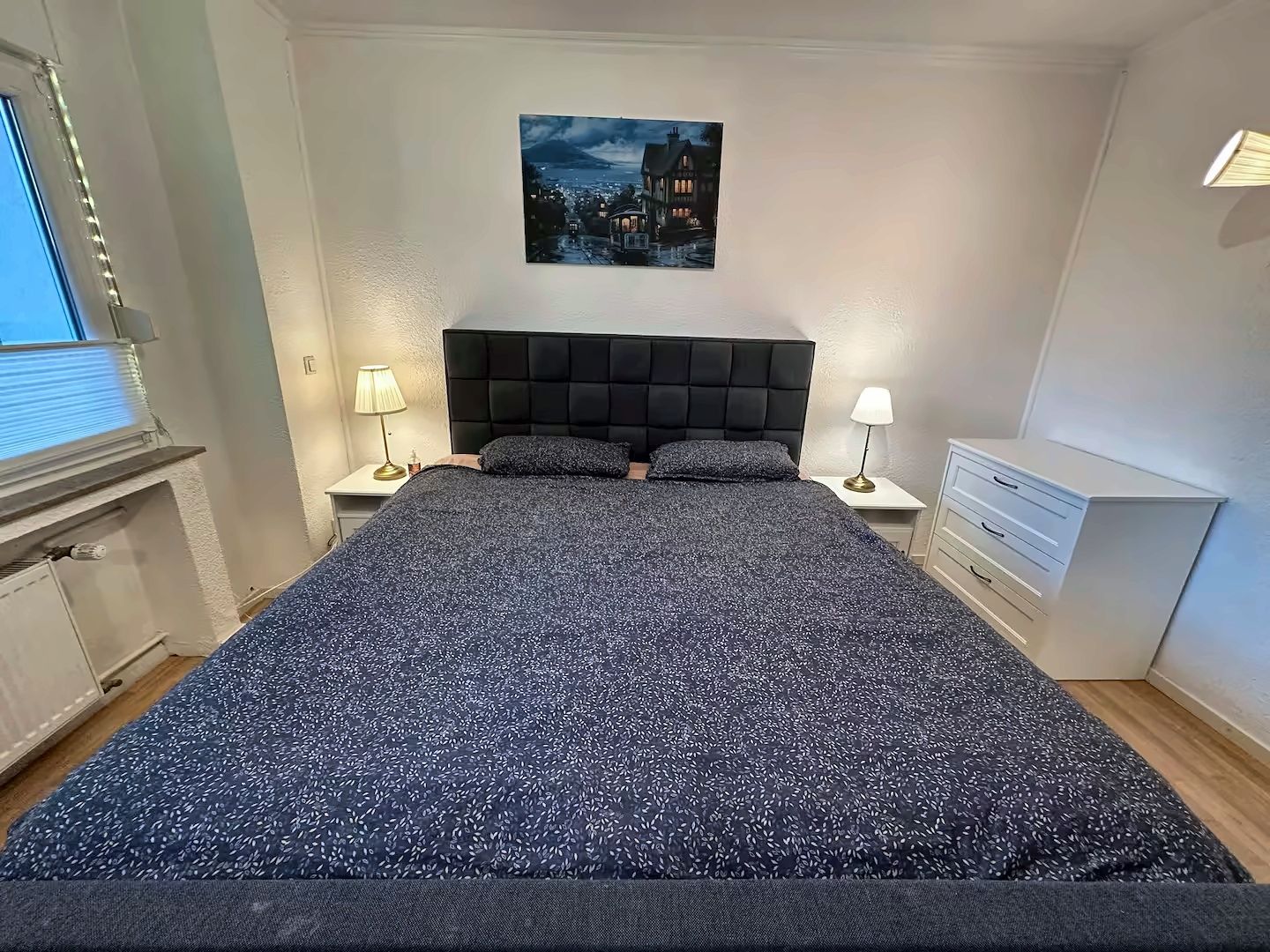 Ein Schlafzimmer mit einem großen Bett, zwei Nachttischen, einem Wandbild und einem Fenster mit Jalousien.