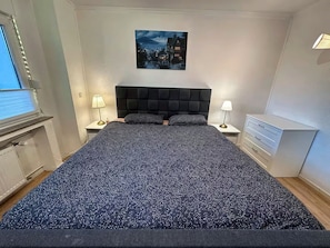A bedroom with a large bed, two bedside tables, a nightstand, a wall-mounted painting, and a window with blinds. - TOP Appartment Nähe Schalke Stadium, Veltins Arena ist 10 min zu Fuß Entfernt (Gelsenkirchen)