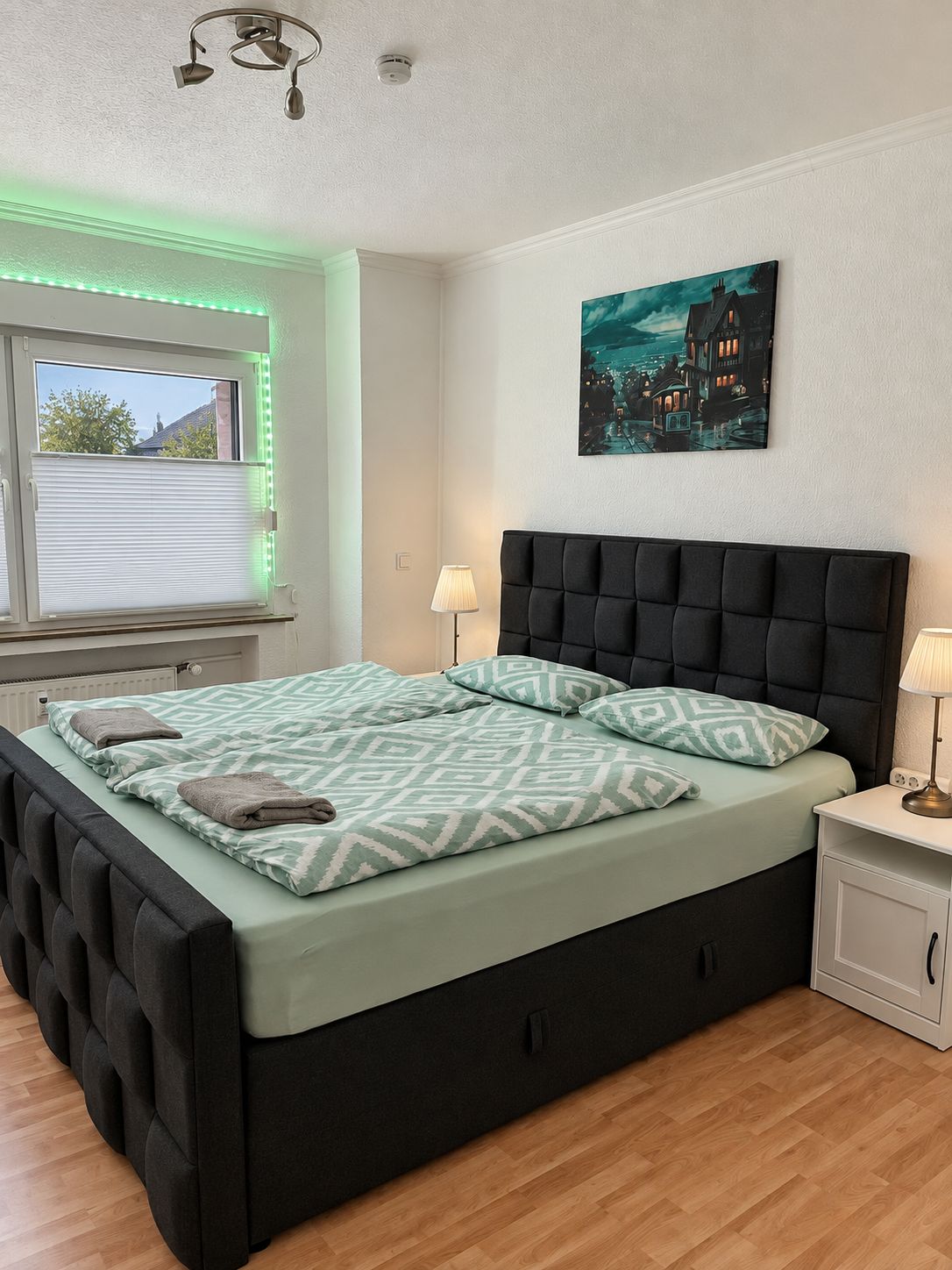 Ein Schlafzimmer mit einem Bett, Nachttischlampen, einem Fenster mit Jalousien, einem Gemälde an der Wand und einer Deckenleuchte.