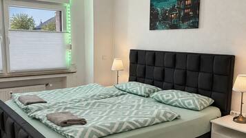 Ein Schlafzimmer mit einem Bett, Nachttischlampen, einem Fenster mit Jalousien, einem Gemälde an der Wand und einer Deckenleuchte.