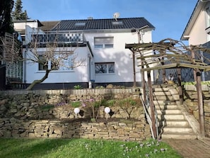 A residential property with a stone retaining wall, a wooden pergola, and a staircase leading to a house with a tiled roof.